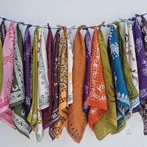 Könnte beinhalten: Eine Sammlung bunter Bandanas, die an einer blauen Schnur vor einer weißen Wand hängen. Die Bandanas weisen verschiedene Muster und Farben auf, darunter Grün, Rosa, Lila, Orange und Blau. Jedes Bandana ist oben verknotet.