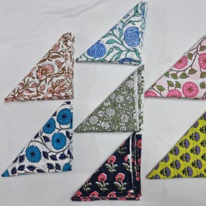 Może przedstawiać: Kolekcja siedmiu złożonych serwetek z tkaniny z kwiatowymi wzorami. Serwetki mają różne kolory, w tym różowy, niebieski, zielony i żółty, z białym tłem. Każda serwetka jest złożona w kształt trójkąta.