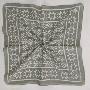 Könnte beinhalten: Ein salbeigrünes Baumwollbandana mit einem weißen floralen und geometrischen Muster. Das Bandana ist quadratisch gefaltet.