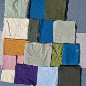 May include: An assortment of folded fabric squares in various colors, including shades of green, blue, yellow, pink, and purple. The fabrics are arranged on a tiled surface, showcasing a variety of textures and hues.