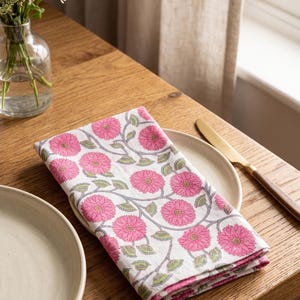 Könnte beinhalten: Gefaltete weiße Serviette mit rosa Blumenmuster auf einem Holztisch. Das Muster zeigt rosa Blüten und grüne Blätter. Ein Teller und ein Messer sind ebenfalls zu sehen.