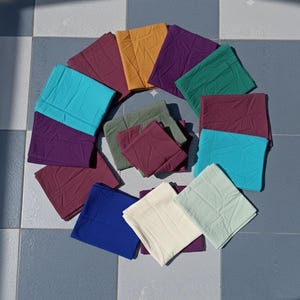 May include: An assortment of folded fabric squares in a variety of colours, such as teal, purple, mustard yellow, and burgundy, arranged in a circular pattern. The squares appear to be made of a soft material, possibly cotton, and are on a tiled surface.