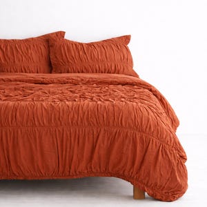 以下が含まれることがあります： テクスチャ加工とギャザーデザインが施された、錆色の寝具セット。セットには、掛け布団カバーと、お揃いの枕カバー2枚が含まれています。生地は柔らかく、しわのある外観です。ベッドは白い背景に置かれています。