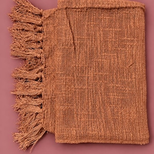 May include: A rectangular, terracotta-colored woven throw blanket featuring a textured, grid-like pattern throughout the fabric. The left edge of the blanket is finished with a row of long, matching terracotta-colored fringe tassels.