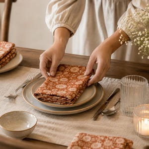 Könnte beinhalten: Eine gefaltete quadratische Stoffserviette mit einem rostorangen Hintergrund und einem sich wiederholenden weißen Blumenmuster. Die Serviette liegt auf einem Stapel aus zwei neutralfarbenen Keramiktellern, ergänzt durch eine Gabel, einen Löffel, ein Messer und ein Glas.