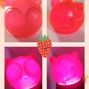 May include: Four images of a cat-shaped night light in a collage. The light is a bright pink, with large, round eyes and small ears. The text "Strawpurry" is at the top. A cartoon strawberry is also present in the image.
