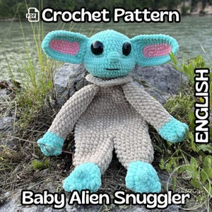 Puede incluir: Una muñeca Baby Alien Snuggler de ganchillo en tonos turquesa, gris y rosa. La muñeca tiene orejas grandes con detalles internos rosas, ojos negros y un aspecto suave y texturizado. La imagen incluye el texto "Crochet Pattern" y "Baby Alien Snuggler".