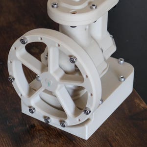 Könnte beinhalten: Ein weißes mechanisches Gerät mit einem kreisförmigen Rad und mehreren Bolzen. Das Gerät hat eine rechteckige Basis und eine Reihe von verbundenen Komponenten. Das Rad hat eine sternförmige Mitte. Das Gerät befindet sich auf einer Holzoberfläche.