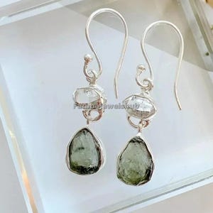 May include: A pair of silver earrings with a clear, faceted top stone and a teardrop-shaped, olive-green bottom stone. The earrings have a simple hook design and are displayed on a clear surface. The brand name "FatimaJewelshub" is visible.