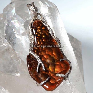 May include: A pendant featuring a polished, reddish-brown gemstone with a unique, textured pattern. The stone is encased in a silver-colored metal frame with a branch-like design. The pendant is displayed against a clear, crystalline background.
