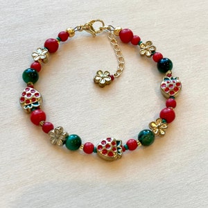 May include: A red and green beaded bracelet with gold accents. The bracelet features small strawberry charms and flower charms.
