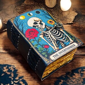 May include: A tarot card-themed journal with a blue cover featuring a skeleton, rose, and the text "THE OVERTHINKER." The journal has a black spine and gold-edged pages. The background is a wooden surface with decorative elements.