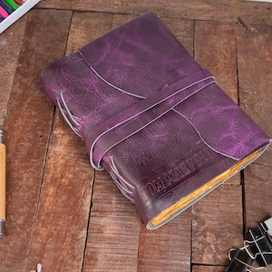 May include: A purple leather journal with a strap closure. The journal has the embossed text "TOM MARVOLO" on the cover. The journal is on a wooden surface.