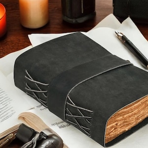 Pu&ograve; includere: Un diario in pelle nera con un'ampia cinghia avvolgente e cuciture incrociate decorative lungo il dorso. Il diario presenta pagine spesse con bordi irregolari e un aspetto invecchiato color marrone chiaro, in contrasto con la copertina scura.