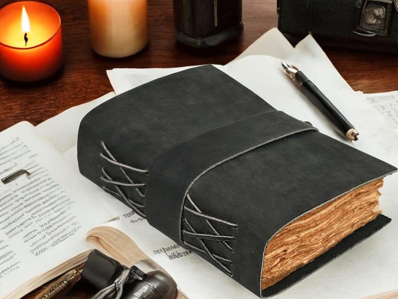 Pu&ograve; includere: Un diario in pelle nera con un'ampia cinghia avvolgente e cuciture incrociate decorative lungo il dorso. Il diario presenta pagine spesse con bordi irregolari e un aspetto invecchiato color marrone chiaro, in contrasto con la copertina scura.