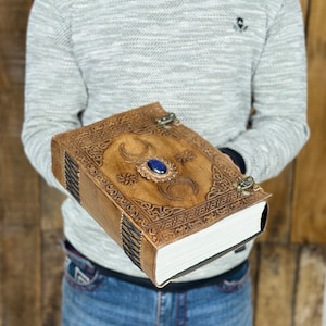 Op de afbeelding: Een bruin leren dagboek met een drievoudig maandesign en een blauwe edelsteen. Het dagboek heeft een decoratieve sluiting en wordt door een persoon vastgehouden.