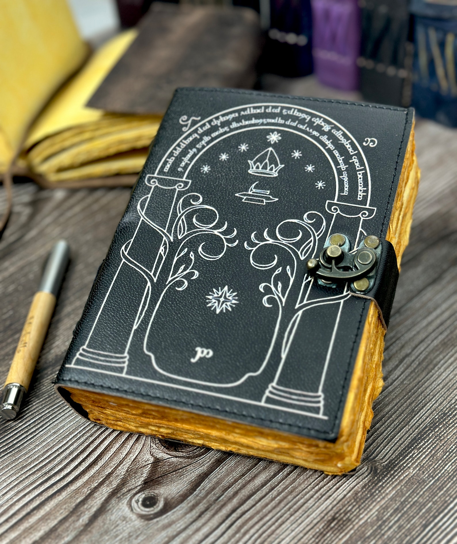 A black leather-bound journal with a metal clasp. The cover is a white outline design of a landmark door in 'The Lord of the Rings'.