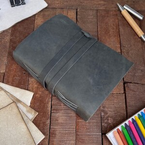 May include: A dark gray leather-bound journal with a stitched spine and three leather straps. The journal is lying on a wooden surface.