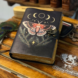 Puede incluir: Un diario de cuero negro con un diseño de luna y estrellas, que presenta una gran polilla con alas rojas y blancas. El diario tiene un cierre de metal y una borla decorativa.