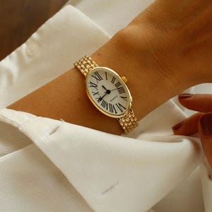May include: Gold-toned wristwatch with an oval face and Roman numeral hour markers. The watch has a gold band and a white watch face. The watch is on a wrist, with a white shirt in the background.