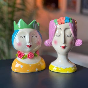May include: Two ceramic face planters. One has a green crown, blue hair, and a yellow polka-dot dress. The other has pink hair, a floral headband, and a yellow polka-dot dress. Both have closed eyes and red lips.