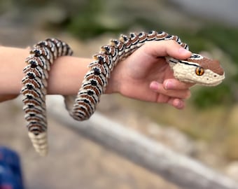 May include: A detailed, realistic toy snake in shades of brown, white, and black is wrapped around an arm. The snake's head is a lighter tan color with a brown top and yellow eyes. The snake's body has a repeating pattern.