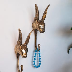 May include: Two gold rabbit head hooks mounted on a white wall. A blue beaded necklace hangs from one of the hooks.