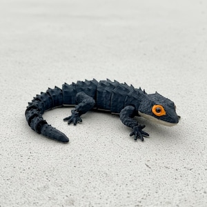 May include: A detailed, black lizard figurine with a segmented body and tail. The lizard has orange eyes and is posed on a light-colored surface. The figurine is a reptile-themed collectible.