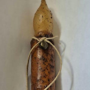 May include: A rustic-style candle with a textured brown body and a light-colored, rounded top. A small metal bell is tied around the candle with twine. The candle has a natural, handcrafted appearance.