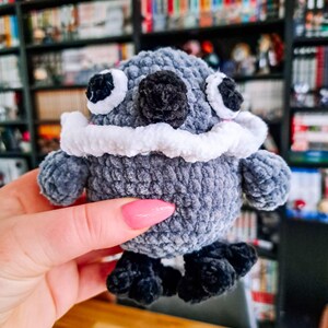 May include: A handmade grey plush creature with black eyes, a black nose, and white trim around the face. The creature has small black feet. The plush toy is a unique, handcrafted item, held in a person's hand.