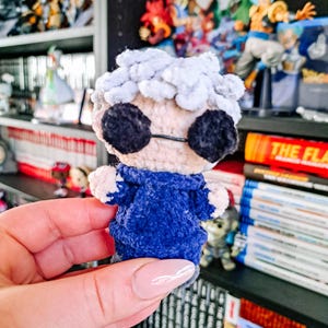 Pode incluir: Boneca de crochê feita à mão com jaqueta azul, óculos pretos e cabelo cinza. A boneca é segurada em uma mão, com uma estante cheia de estatuetas e livros ao fundo. O rosto da boneca é bege claro e os óculos são redondos.