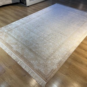 May include: A cream-colored rug with a subtle floral pattern and a fringed border. The rug has a gradient effect, with the center being lighter than the edges.