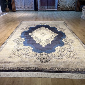 May include: A large rectangular rug with a central medallion design in shades of blue and cream. The rug features intricate floral patterns and a dark blue border with fringe along the edges. The rug is displayed on a hardwood floor.