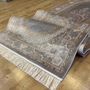 May include: A gray and beige patterned rug with a white fringe. The rug has a traditional design with intricate details.