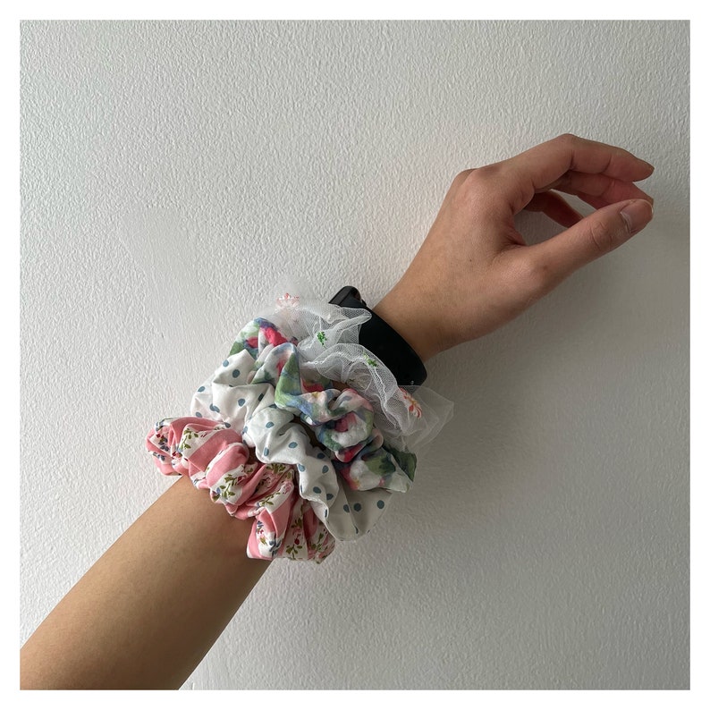 May include: A set of three hair scrunchies in floral and polka dot patterns. The scrunchies are stacked on a person's wrist.