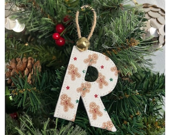 May include: A fabric letter R ornament featuring a repeating pattern of brown gingerbread men, small red stars, and tiny green stars on a cream background, topped with a gold jingle bell and a natural jute twine hanging loop.