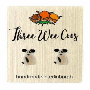 Puede incluir: Pendientes con forma de perro sobre una tarjeta crema. Los pendientes son blancos con manchas y orejas negras. La tarjeta tiene el texto "Three Wee Coos" y "handmade in edinburgh", con una ilustración de tres vacas de las Highlands.