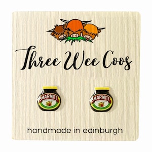 May include: A pair of earrings featuring a cartoon image of a jar of Marmite, a popular British yeast extract spread. The earrings are handmade in Edinburgh, Scotland. The card also features a cartoon image of three Highland cows with the text "Three Wee Coo's".
