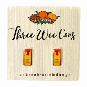 May include: A pair of earrings featuring miniature cans of Tennent's Lager, a Scottish beer. The earrings are displayed on a wooden card with the text "Three Wee Coo's" and "handmade in Edinburgh".