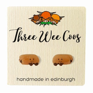 Könnte beinhalten: Zwei braune, lächelnde, cartoonartige Würstchen-Ohrringe mit dunkelbraunen Füßen. Die Ohrringe sind auf einer cremefarbenen Karte mit dem Text "Three Wee Coos" und "handmade in edinburgh" abgebildet. Oben ist ein Cartoon von drei Highland-Kühen.