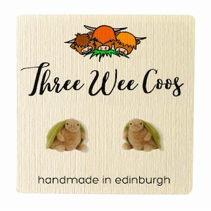 Könnte beinhalten: Eine cremefarbene Karte mit dem Text "Three Wee Coos" und einer Illustration von drei Highland-Kühen. Darunter befinden sich zwei kleine, filzartige Kreaturen in Beige mit grünen Akzenten. Der Text "handmade in edinburgh" steht unten.