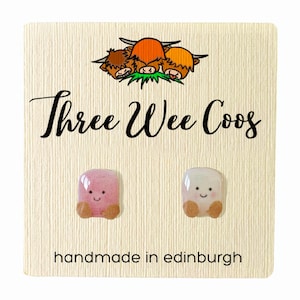 May include: A pair of pink and white ceramic earrings shaped like Highland Cows with smiling faces. The earrings are handmade in Edinburgh and are displayed on a card with the text "Three Wee Coo's".