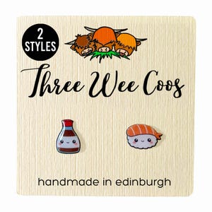 May include: A card with a brown background and the text "Three Wee Coo's" in black cursive font. The card also features a cartoon image of three Highland cows, a cartoon image of a bottle of soy sauce with a smiling face, and a cartoon image of a piece of sushi with a smiling face. The card also has the text "2 Styles" in a black circle and the text "handmade in edinburgh" in black text.