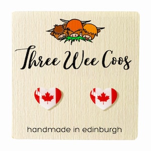 May include: A pair of heart-shaped earrings with the Canadian flag design, displayed on a cream card. The card reads "Three Wee Coos" and "handmade in edinburgh", with a cartoon of three Highland cows.