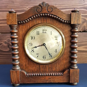 May include: Antique wooden clock with a round, white face and black hands. The clock has Roman numeral markings and is set in a dark wood frame with intricate carvings.