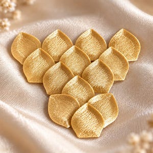 Könnte beinhalten: Eine Ansammlung von zwölf goldfarbenen, blütenblattförmigen Stickapplikationen, die in einem Dreiecksmuster angeordnet sind. Jedes einzelne Teil hat eine spitze Form und eine texturierte Oberfläche mit feinen Metallfäden.