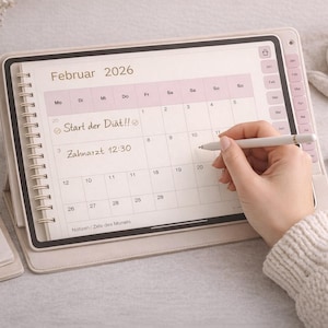 Könnte beinhalten: Ein digitaler Kalender für Februar 2026 ist geöffnet, mit einer Hand, die einen Stift hält, um auf den Bildschirm zu schreiben. Der Kalender befindet sich in einem hellfarbigen Gehäuse, mit einem Spiralnotizbuch und einem Stift in der Nähe. Der Text "Start der Diät!!" ist sichtbar.