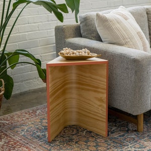 May include: A wooden triangular side table with a light wood finish and a pink trim. The table is sitting on a rug with a floral pattern and has a small bowl of white beads on top.