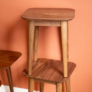 May include: Two wooden side tables with rounded corners and tapered legs are stacked. The tables are a warm brown color and appear to be made of hardwood. The background is a solid orange-red color.