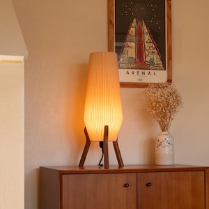 A mid-century modern style table lamp with a cream-colored, ribbed shade. The lamp sits on a dark wood tripod base. The lamp is turned on, emitting a warm glow. A framed poster and a vase with dried flowers are in the background.